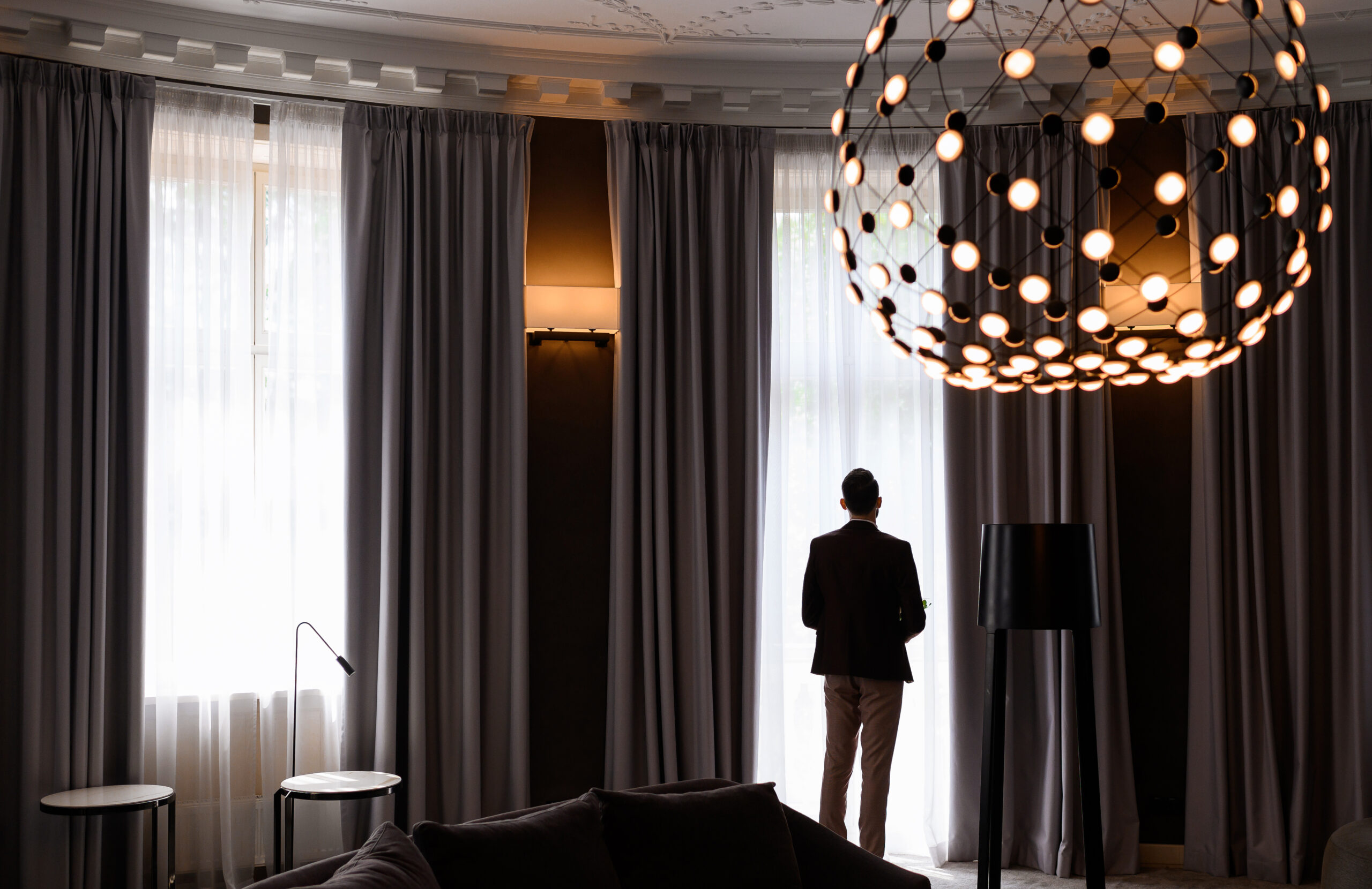 Back view of groom in wedding black suit standing in luxury spacious apartment with designer large chandelier, and looking at window while waiting on wedding ceremony. from freepick